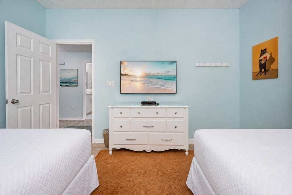Bright guest bedroom featuring two double beds, a dresser with a TV, and cheerful beach artwork on soft blue walls