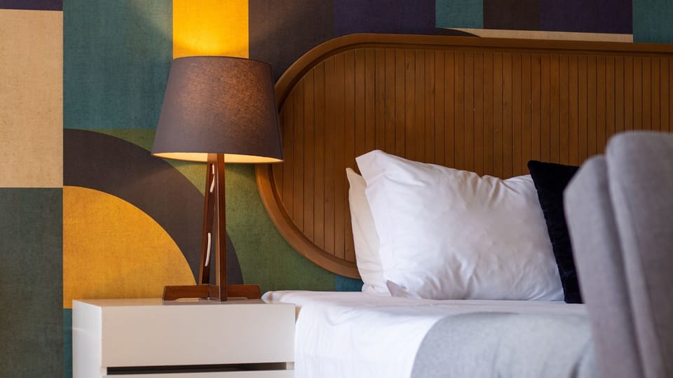 Drift off to a peaceful sleep in this inviting bed, where the warm glow of the bedside lamp creates a tranquil ambiance against the vibrant, artistic wallpaper. The elegant wooden headboard adds a touch of natural sophistication to your private retreat.