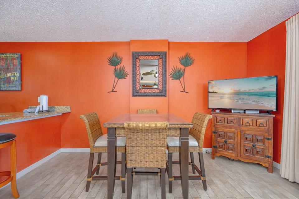 The dining area features a glass-top table with wicker chairs, a large TV, and a stunning beach view through sliding doors