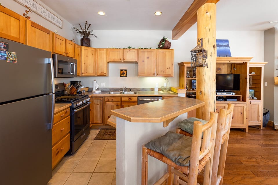 A cozy kitchen and dining area with wooden cabinets, stainless steel appliances, a bar counter with two stools, and a living room area in the background.