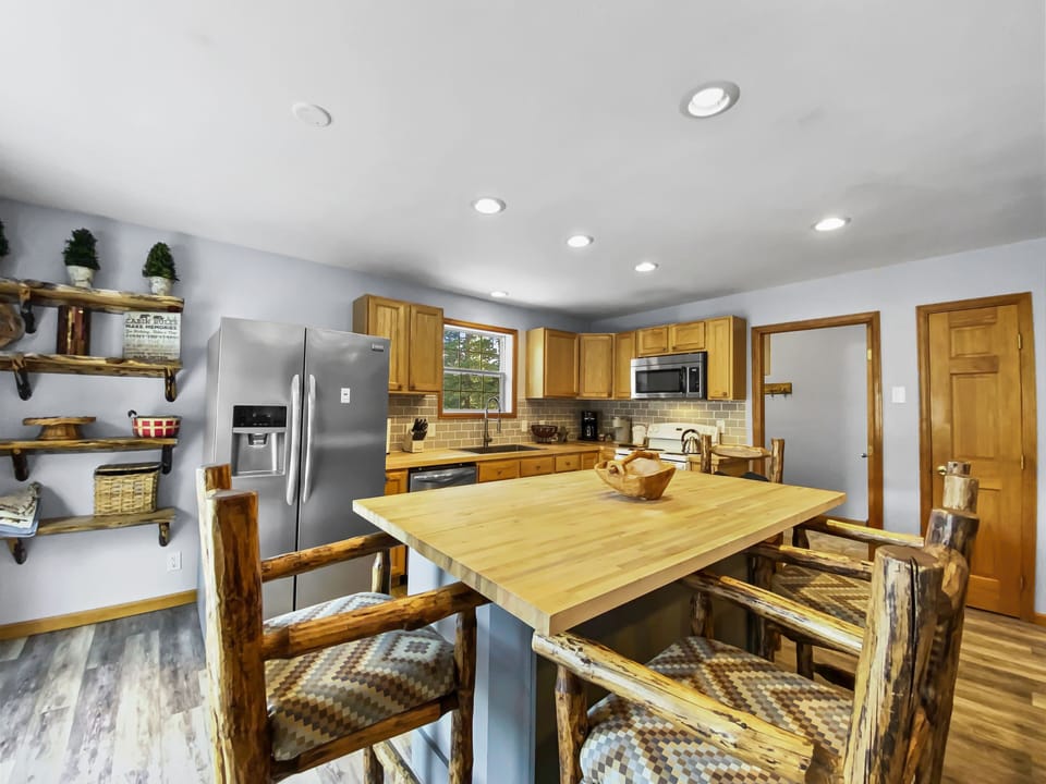 Kitchen with wooden cabinetry, stainless steel appliances, an island with a wooden top, and four log-frame barstools. Shelves hold potted plants, baskets, and kitchen items. Ceiling lights are on.