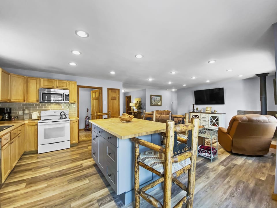 A spacious, open-concept kitchen and living area with wooden cabinetry, a center island with rustic chairs, a white stove, and a microwave. The living area features cozy seating and a wall-mounted TV.