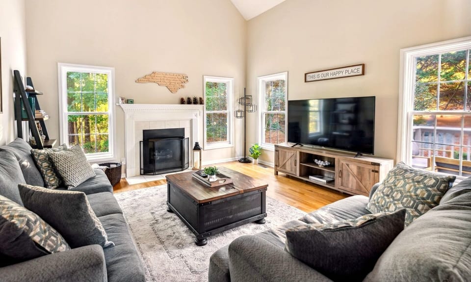 Gather in the light-filled living room with vaulted ceilings, cozy seating, and a central fireplace overlooking peaceful wooded views.