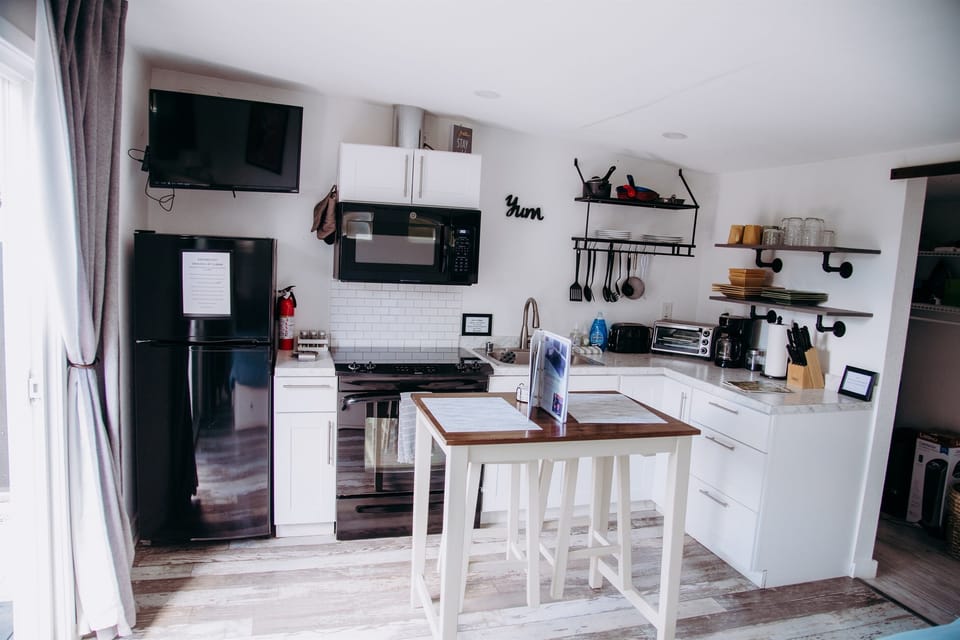 This modern studio kitchenette features clean white cabinetry, a marble-inspired countertop, and a versatile central island. Outfitted with a black refrigerator, gas range, and wall-mounted TV, it offers a stylish space.