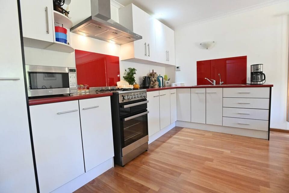 This modern kitchen features sleek white cabinetry with chrome handles, striking red countertops and backsplash, and stainless steel appliances including a gas stove and microwave. The space is bright and functional, with ample storage and a clean, contem