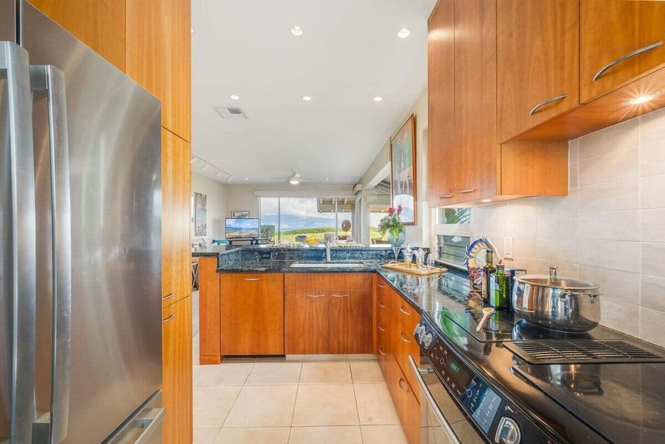 Modern kitchen with stainless steel appliances, warm wood cabinetry, granite countertops, and an ocean view through large windows.