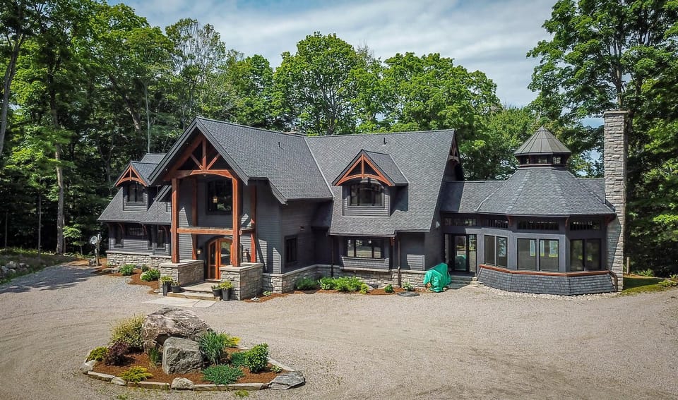 Front exterior of the cottage showcases dark gray shingles, stone accents, and wooden beams framing the covered entryway.