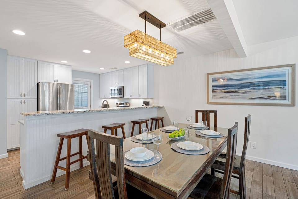 Just beyond, the kitchen features a long breakfast bar with ample seating, stainless steel appliances, and clean, coastal finishes.