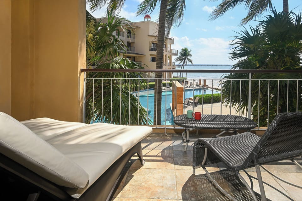 Step onto your private balcony and unwind on cushioned loungers overlooking the sparkling pool, swaying palms, and glittering sea beyond—your perfect spot to soak up the coastal breeze.