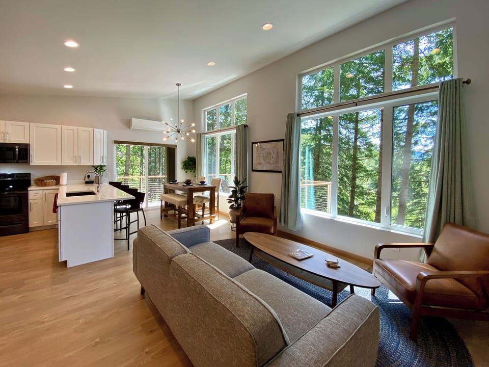 The whole picture — open-plan living, dining, and kitchen wrapped in floor-to-ceiling forest views on every side