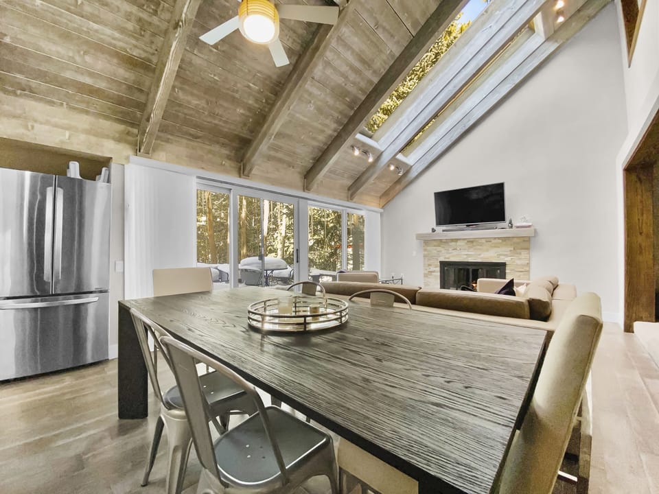 A modern, open-concept living and dining area featuring a wooden table with metal chairs, a sofa, a fireplace, a wall-mounted TV, large windows, and a stainless steel refrigerator.