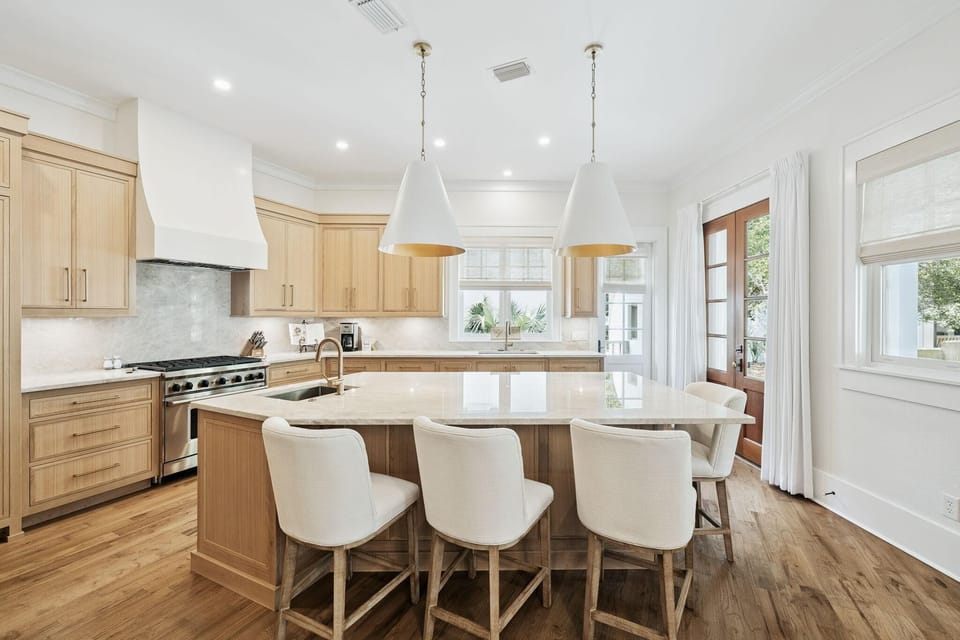 Large kitchen island with marble countertop, four white upholstered stools, and light wood cabinetry.