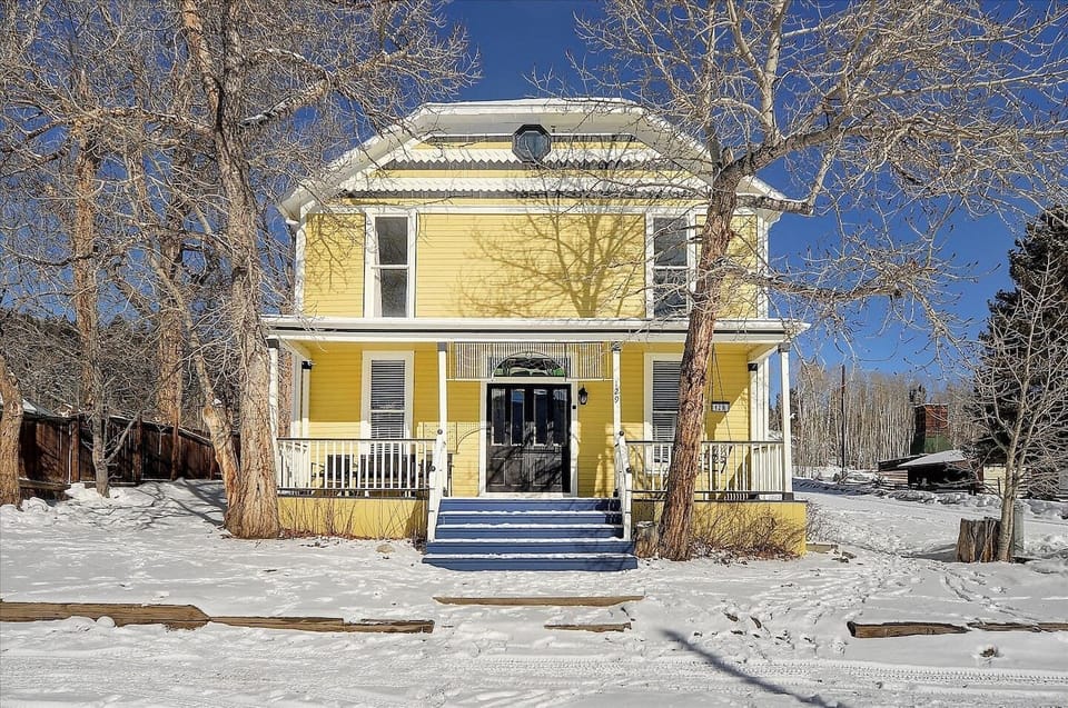 Charming yellow Victorian style home with black front door, wraparound porch, and snowy winter curb appeal.