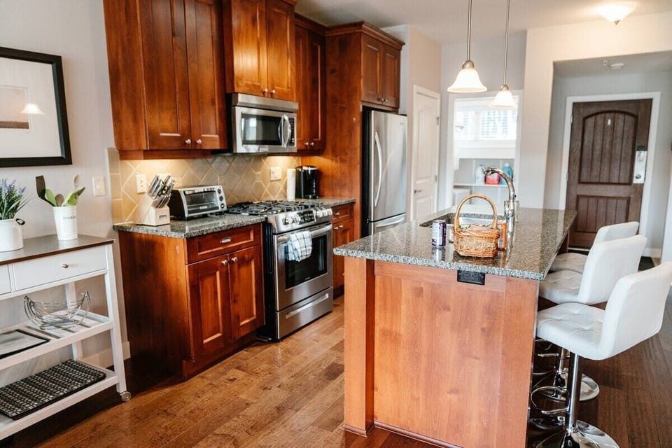 This inviting kitchen features rich wooden cabinets, stainless steel appliances, and a spacious granite island with seating. Pendant lights and tasteful decor, along with warm hardwood floors, create a cozy atmosphere.
