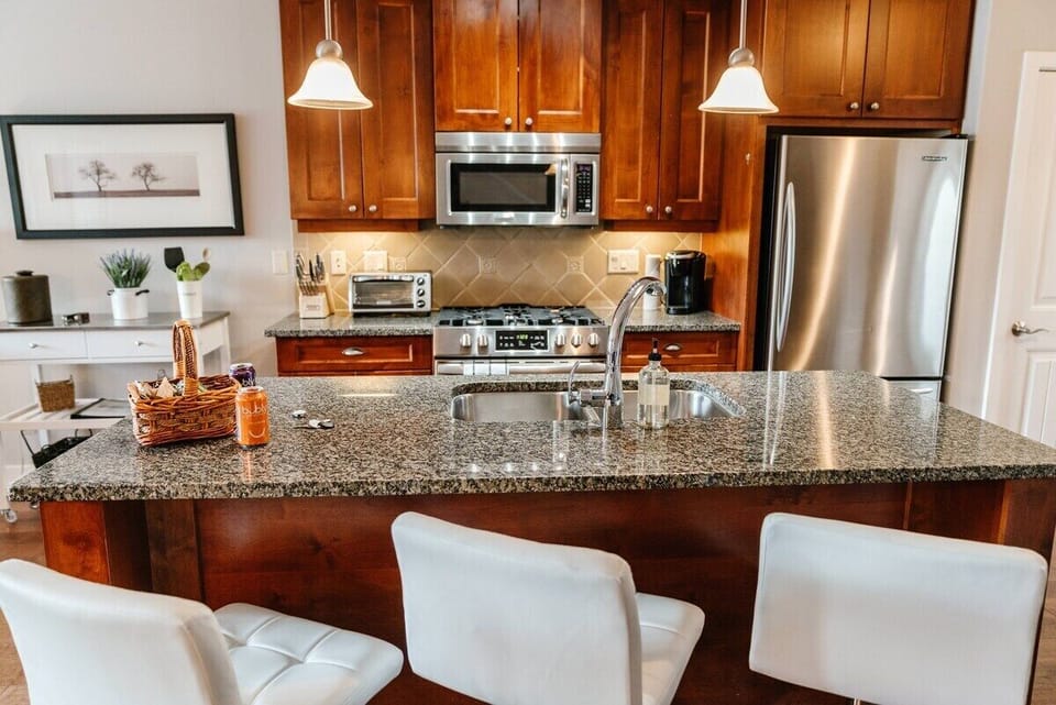 This modern kitchen features a spacious island with a speckled granite countertop and white leather bar stools. Warm wood cabinets, stainless steel appliances, and a stylish mosaic backsplash create a sleek, inviting space.