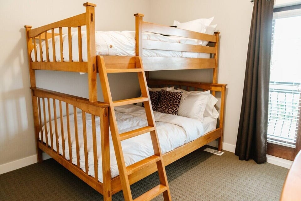 This cozy bedroom features a light brown wooden bunk bed with traditional design and a ladder for easy access to the top bunk. Both bunks are made with white sheets and pillows, accented by leopard print pillows on the bottom bunk.
