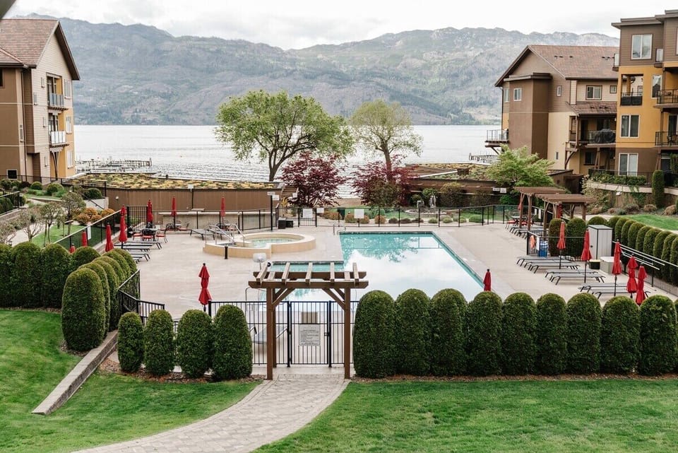 Relax in style at this resort-like pool area, featuring a serene lake view, red umbrellas, and cozy lounge chairs. A wooden pergola offers shade, while rolling mountains set a peaceful backdrop.