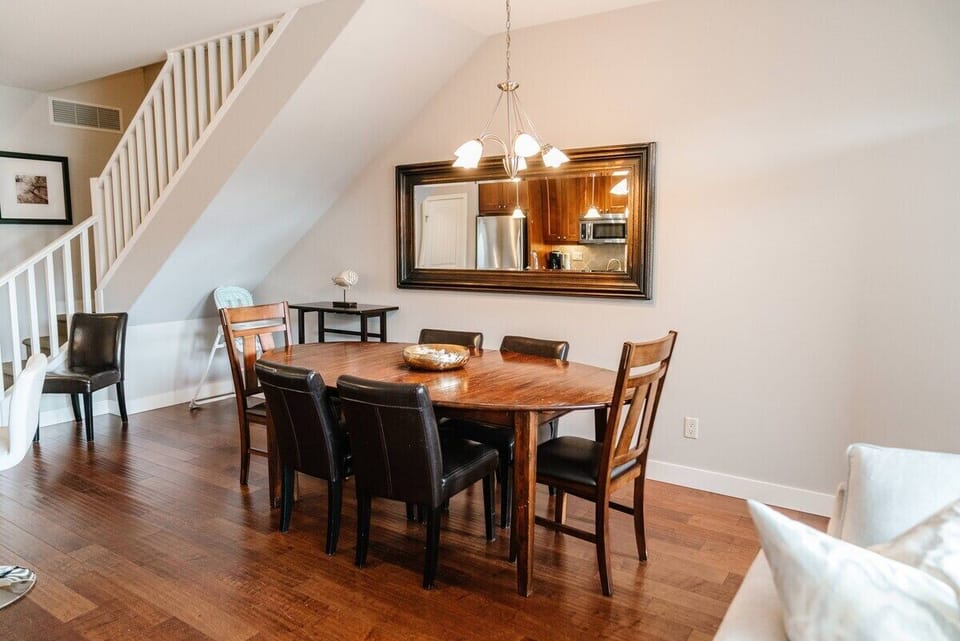 This stylish dining area features an oval wooden table with six leather chairs, set with a charming centerpiece. A large mirror reflects the kitchen, while natural light and hardwood floors enhance the space.