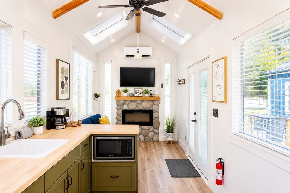 Bright open-concept tiny home kitchen and living area with vaulted ceilings, skylights, modern fireplace, Smart TV, and natural light throughout the cozy space.