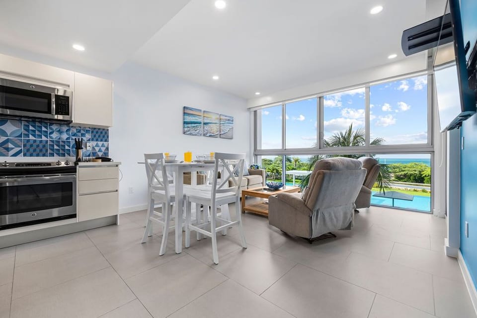 Step into your bright, open living space where the ocean beckons through floor-to-ceiling windows. Relax in the cozy armchair, share meals at your dining table, and enjoy the modern kitchen ready for your culinary adventures.