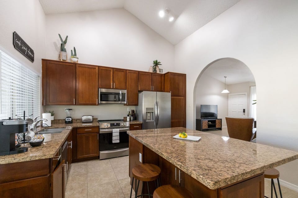 Modern kitchen with stainless steel appliances, wooden cabinets, and granite countertops. Decorative plants and small appliances on countertops. Archway leads to a living room with a TV and dining area.