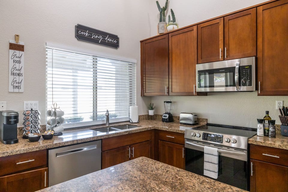 A modern kitchen with wooden cabinets, granite countertops, a stainless steel stove, microwave, and dishwasher. A window with blinds is above the sink. Decor items and small appliances are on the counter.