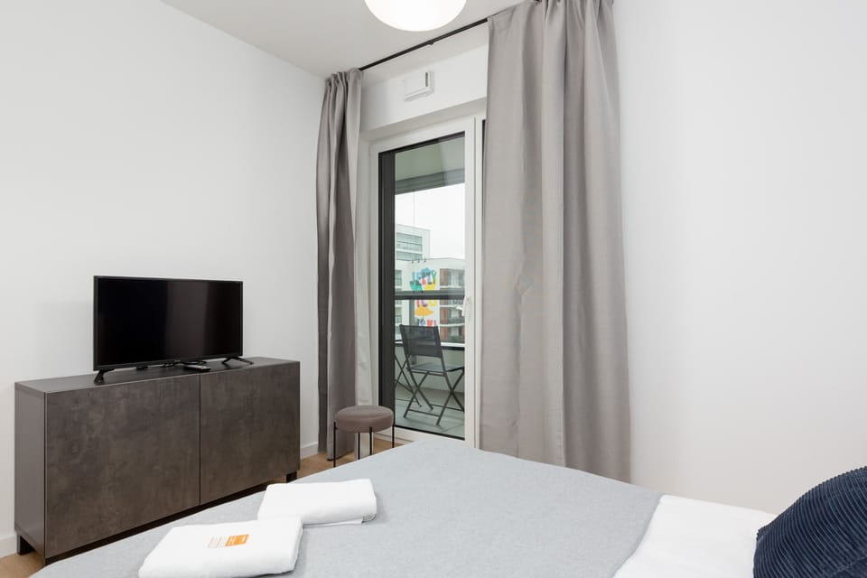 Another view of the bedroom with a double bed with a grey bedspread, Opposite the bed is a white cupboard on which stands a flat-screen TV.