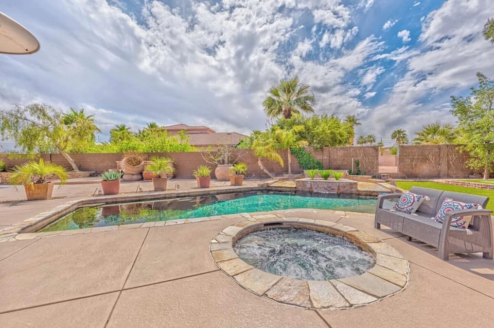 A backyard patio features a round hot tub in the foreground and a rectangular swimming pool surrounded by potted plants, with palm trees