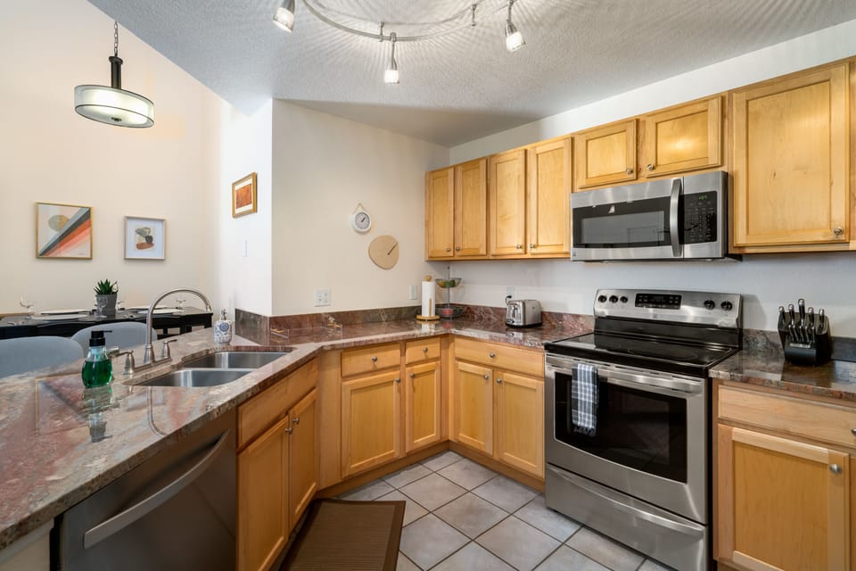 A modern kitchen with wooden cabinets, stainless steel appliances, granite countertops, a double sink, and tiled flooring. Adjacent dining area is visible in the background.