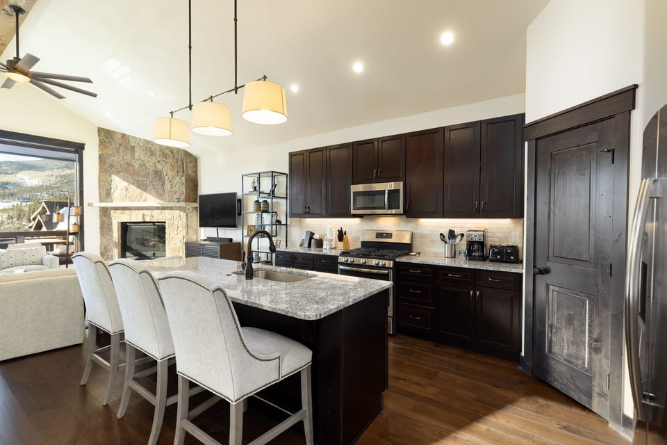 A modern kitchen with dark wood cabinets, stainless steel appliances, a large island with stone countertop and three upholstered bar stools. An open living area with a fireplace is in the background.
