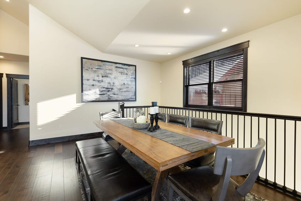 A dining area with a wooden table, black cushioned benches, and chairs. There is abstract artwork on the wall, a large window with blinds, and a railing separating it from the adjacent room.
