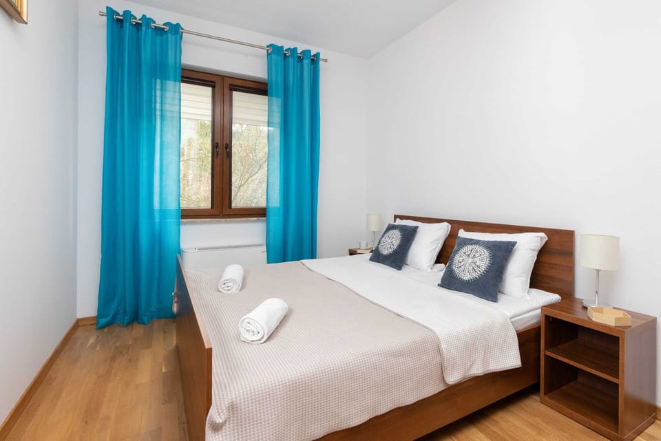 A bedroom featuring a large bed with a white bedspread and decorative blue pillows. Bright blue curtains hang by the window, and there are bedside tables with small lamps.