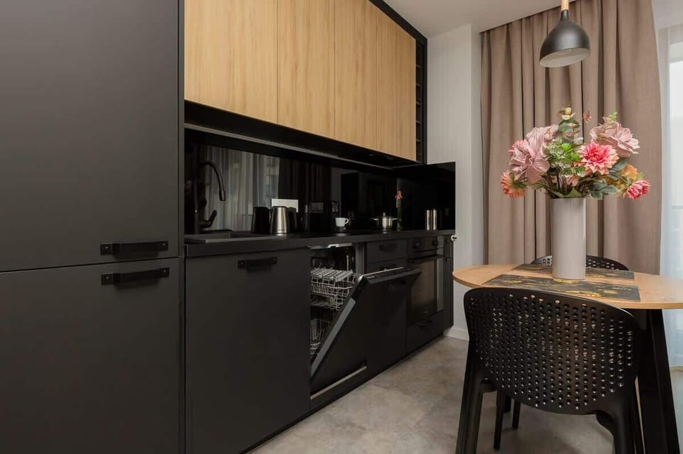 A sleek kitchen with black cabinets, modern appliances, and a wooden backsplash. A small dining table is set with flowers as a centerpiece.

