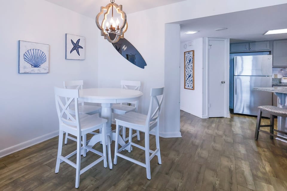 Bright dining area featuring a round white table with four chairs, coastal artwork, and a view into the kitchen