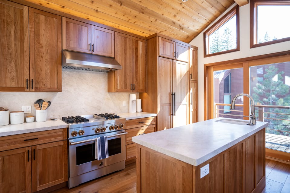 Showing new (2021) JennAir gas oven/range, large refrigerator/freezer, natural stone counter, island w/sink, cherry cabinets w/pull out drawers, and large sliding glass door leading to deck.