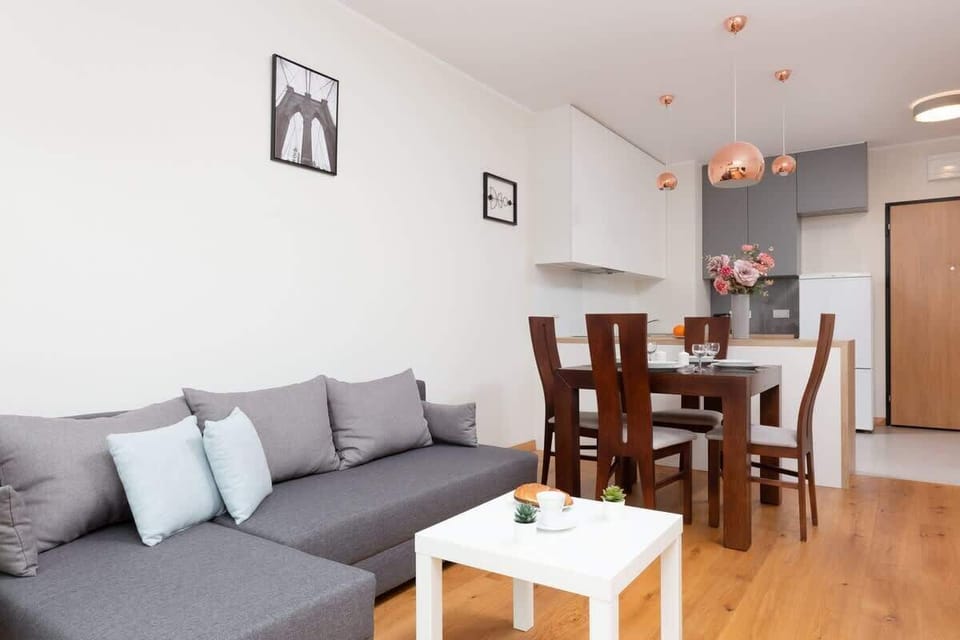 An open-concept living and dining area with a gray sofa, a dining table with four chairs, and light wooden floors. The room has a welcoming and airy ambiance.