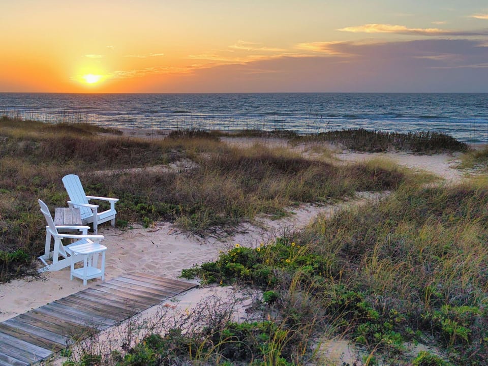 Beachfront boardwalk with seating overlooking the ocean during sunrise, featuring natural dune vegetation and sandy pathways.