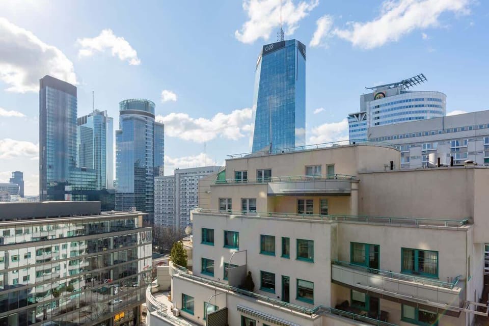 A view from the balcony or window showing a cityscape with modern buildings and skyscrapers. The sky is bright and clear, adding to the urban charm.