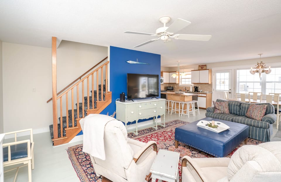 Bright open living room with striped sofa, two armchairs, large blue ottoman coffee table, patterned rug, flat-screen TV on console against blue accent wall, staircase, and open kitchen with bar stools and dining area in this Oak Island beach house.