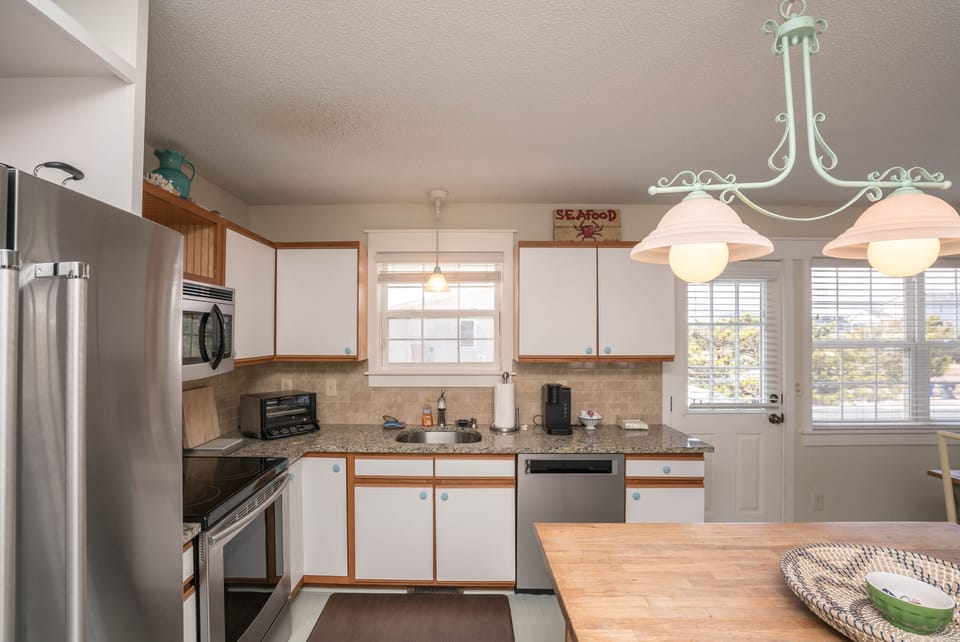 Bright kitchen with stainless refrigerator, stove, microwave, dishwasher, granite countertops, wood island with three stools, pendant lighting, coffee maker, white cabinets, and door leading outside in this Oak Island beach house.