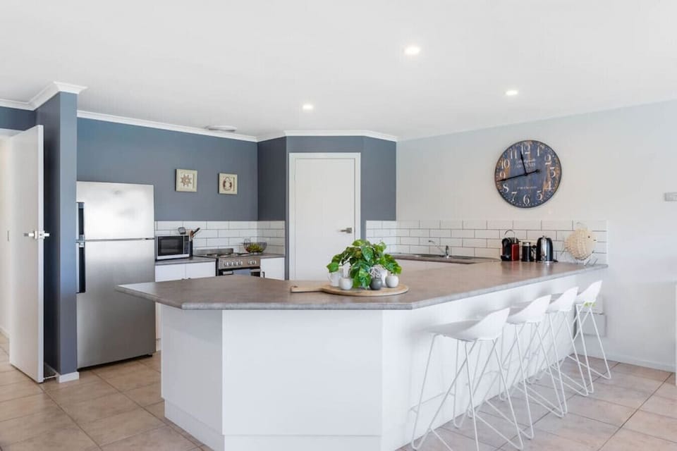 This modern kitchen features a large wraparound countertop with seating for four, white subway tile backsplash, and sleek stainless-steel appliances. Navy blue accent walls and a rustic wall clock add a stylish coastal touch.