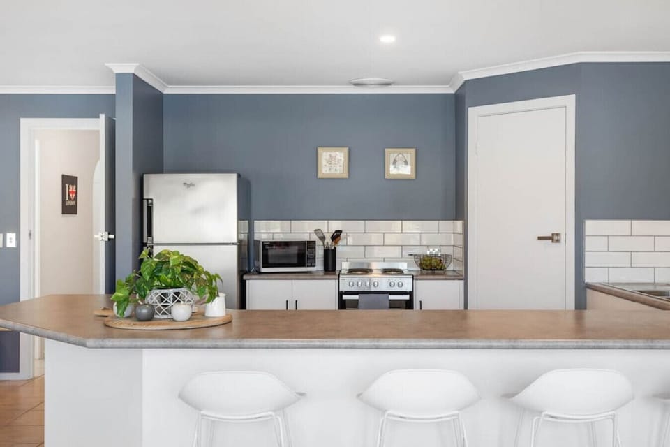 This contemporary kitchen features sleek white cabinetry, a stainless steel fridge, and a compact cooking area with a microwave and stove. The blue-gray walls contrast nicely with white subway tile backsplashes and neutral countertops, while a touch of gr