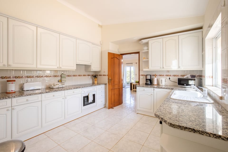 This image shows a bright and functional kitchen with clean white cabinets and plenty of natural light. A large window above the sink provides a nice view of the pink flowering plants outside. The kitchen is spacious and looks very inviting.