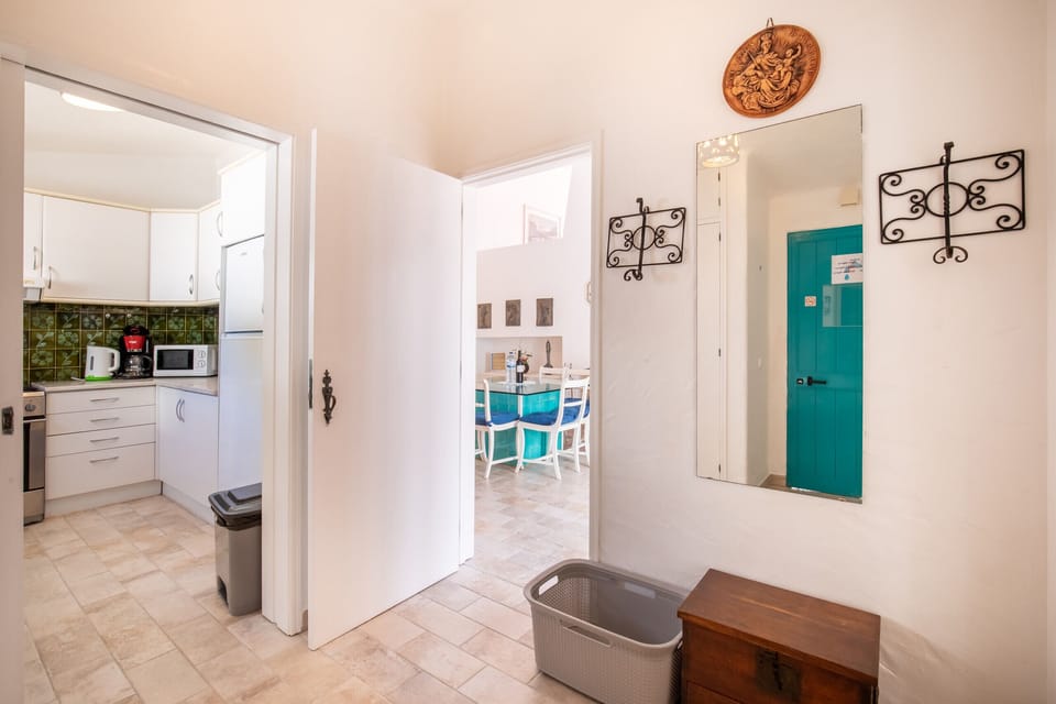This photo shows an interior hallway that provides a direct view into the kitchen and the dining area. Through the open doorway on the left, the kitchen with its white cabinets and green tiled backsplash is visible. In the distance, through another open archway, is the dining area.