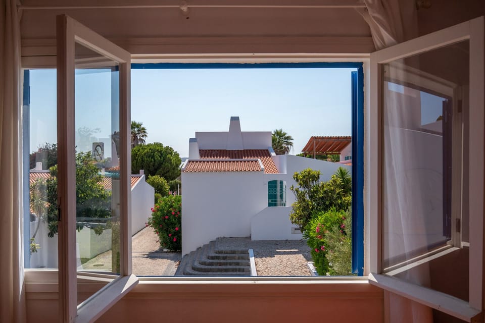 This photo offers a view from an open window towards the outdoor area of the accommodation. One can see the entrance courtyard with the paved path leading to the white buildings with the traditional blue shutters. The surrounding bushes and trees highlight the natural and tranquil setting of the house.