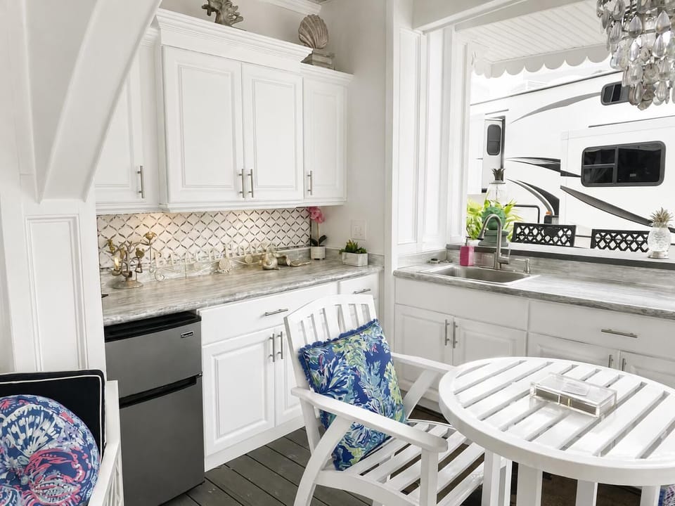 Outdoor kitchen area with marble countertops, white cabinetry, stainless mini fridge, and white slatted table and chairs.