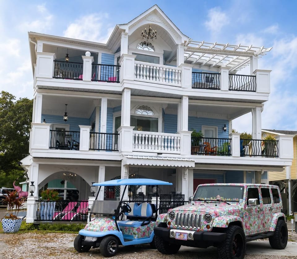 Three-story blue house with white trim, multiple porches with black and white railings, and a floral-patterned Jeep and golf cart parked outside.