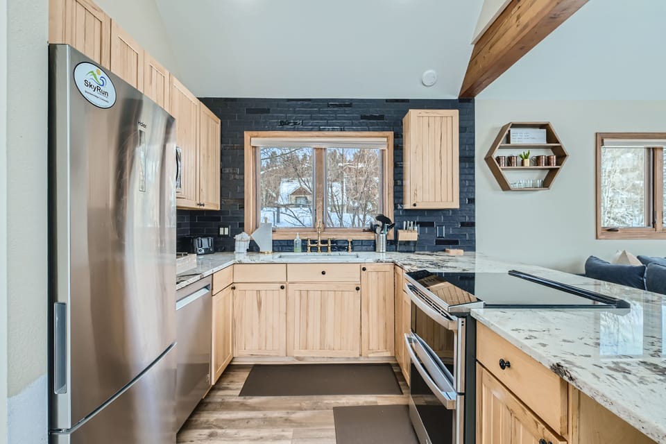A modern kitchen with wooden cabinets, a stainless steel fridge, a double oven, marble countertops, and a central window over the sink. The room features a black tiled backsplash and open shelves.