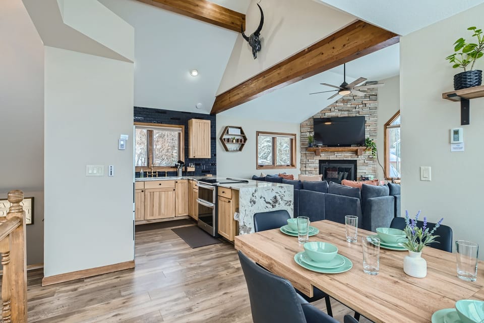 Open-plan living space featuring a dining area with a wooden table set for four, a cozy living area with a fireplace and wall-mounted TV, and a kitchen with wooden cabinets and modern appliances.