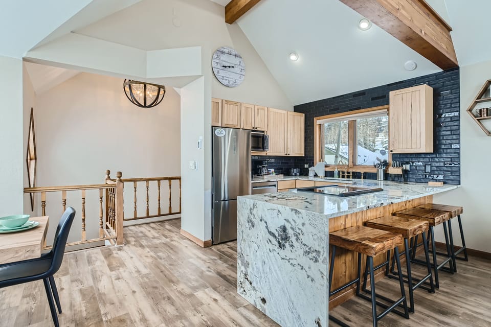 A modern kitchen with wooden cabinetry, stainless steel appliances, and a marble island with bar stools. In the background, a dining area and hallway with a staircase are visible.